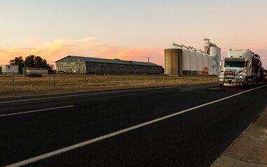 A semitrailer driving past grain silos with pretty sunset in the background