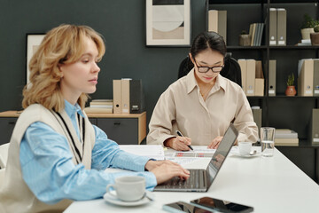 Obraz premium Candid shot of two young businesswomen coworking in open office setting busy with individual tasks and using laptop