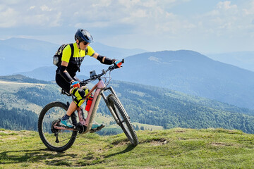 Fototapeta premium Cyclist man riding electric mountain bike outdoors. Male tourist biking along grassy trail in the mountains, wearing helmet and backpack. Concept of sport, active leisure and nature.