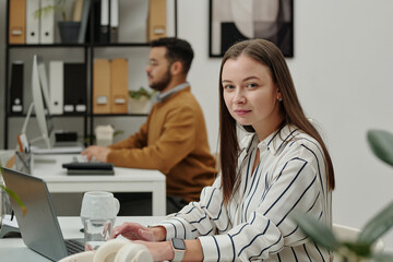 Naklejka premium Portrait of of young woman looking at camera while sitting at workplace in modern office and using laptop copy space 