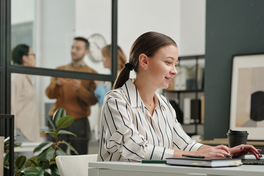 Side view portrait of smiling young woman using computer while sitting at desk and enjoying work in office 