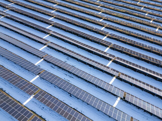 Aerial view of a large solar farm covered in snow, surrounded by a rural landscape with scattered houses and trees. The solar panels contrast against the white ground, showcasing renewable energy in