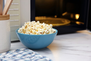 Tasty popcorn in bowl near microwave oven on white marble table, space for text