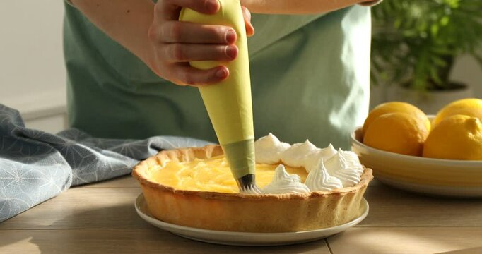 Woman squeezing whipped egg whites onto lemon pie at wooden table, closeup. Making meringue dessert