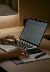 Person using laptop on wooden desk in low light with smartphone and notepad