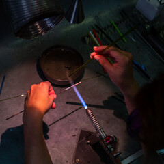 Close-up of a glass artist using a torch to shape molten glass in a workshop. The hands and tools are in focus, highlighting craftsmanship