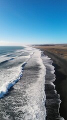 Panoramic view of a black sand beach with foamy waves stretching to the horizon under a clear blue sky