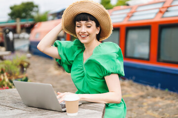 Young woman enjoys outdoor workspace by the canal, wearing a green dress and a straw hat, working...