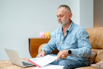 Middle-aged man organizes important paperwork while working on a laptop at home during the day