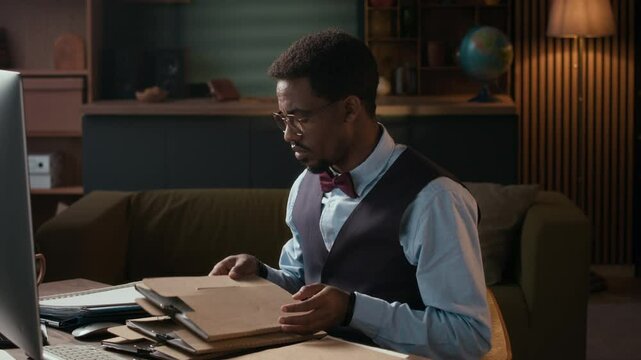 Medium shot of male African American dean in formalwear checking papers and folders sitting at table in home office