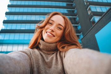 Young woman with long red hair smiles for a selfie outside a modern building on a sunny day