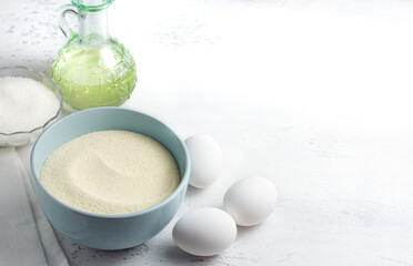 Baking ingredients: light blue bowl with semolina, fresh white eggs, a glass bowl of granulated sugar, and a bottle of vegetable oil on a white kitchen surface