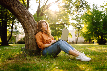 Obraz premium Happy woman with laptop working in a sunny park on a green lawn. Young female freelancer enjoying the weather outdoors. Remote work concept. Active lifestyle.