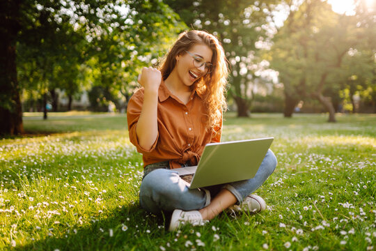 Happy woman with laptop working in a sunny park on a green lawn. Young female freelancer enjoying the weather outdoors. Remote work concept. Active lifestyle.