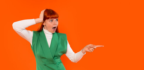 Woman in green vest with red hair gestures in surprise against bright orange background during a lively indoor event