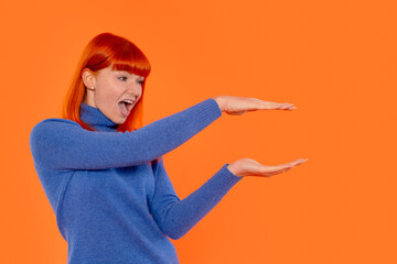 Red-haired woman joyfully gesturing in front of vibrant orange backdrop during indoor portrait...