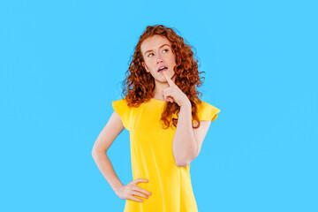 Young woman in yellow dress thoughtfully pondering against bright blue background