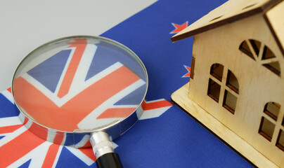 Wooden house model, magnifying glass and New Zealander flag on gray background. Find real estate in...