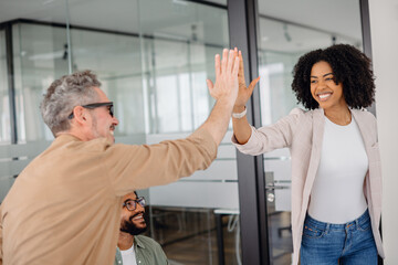 Obraz premium A smiling businesswoman and a senior colleague share a high-five in a collaborative office space. Teamwork, motivation, success, and strong professional relationships in a corporate environment.