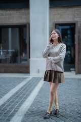 Fototapeta premium Stylish young Asian businesswoman wearing a cardigan and skirt, walking confidently while talking on a smartphone in a bustling city street during the daytime