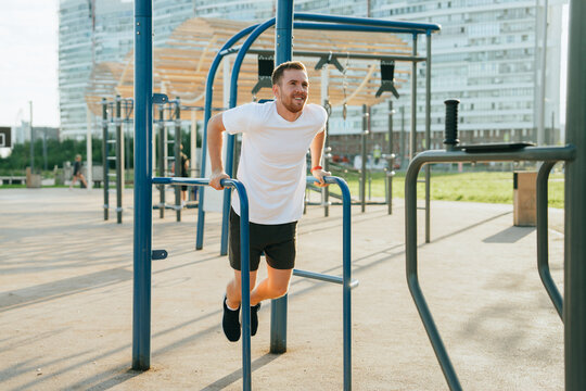 A man exercises energetically on parallel bars in an outdoor urban gym, enjoying a sunny day with a backdrop of modern city buildings.