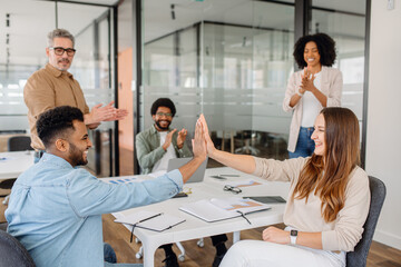 Two young professionals exchange a high-five at a desk, symbolizing teamwork and shared success. Colleagues celebrate an achievement in a modern office space, representing motivation and camaraderie.