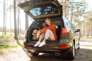 Young woman in stylish clothes relaxing in the trunk of a car. Beautiful woman enjoying sunny...