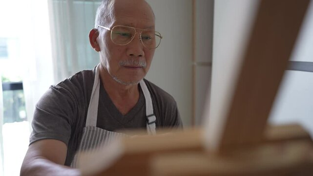 An older asian man, wearing an apron, paints on a canvas at his home during the daytime. Enjoying his artistic hobby in retirement.
