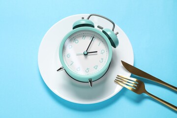 Time for meal. Alarm clock, plate and cutlery on light blue background, flat lay