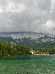 A serene alpine lake with emerald-green water reflects the dense forest. Heavy gray clouds descend...