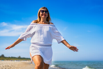 Summertime by sea. Beautiful middle aged woman tourist walking on sandy beach on sunny summer day....