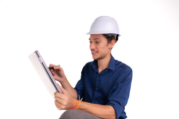 Young architect wearing white helmet, sitting, working with tablet. Isolated on white background.