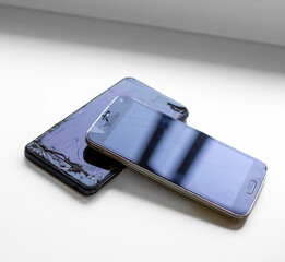 Two damaged smartphones with cracked screens stacked on a white surface, reflecting light. Concept of device repair, damage, and technology issues