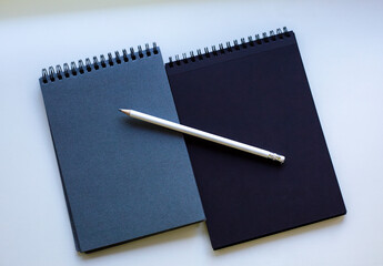Two spiral-bound notebooks, one dark blue and one black, with a white pencil placed diagonally on them, resting on a white surface. Concept of writing