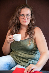 Lovely young woman with long curly hair holding a cup of hot coffee and resting her hand on an open...