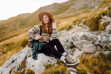 Naklejka premium A woman hiker in a hat and with a backpack enjoys the view of the mountains. Young woman feels freedom on top of a mountain at sunset. The concept of enjoyment, freedom, hiking. Active lifestyle.