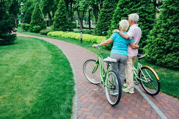 A senior couple takes a break on their bicycles, embracing in a peaceful park surrounded by greenery and walking paths