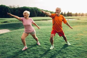 Two seniors perform exercises on a grassy field, enjoying the warm evening sunlight and the natural surroundings