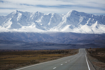 A road stretching into the distance toward the majestic North Chuysky Ridge with snow-capped peaks in the Altai Republic, creating a breathtaking and serene scene.