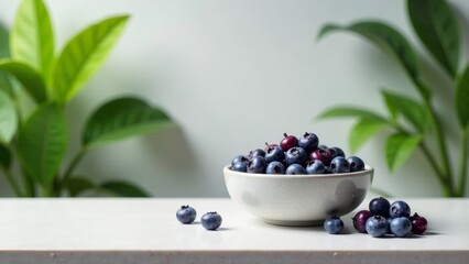 A ceramic bowl filled with ripe blueberries sits on a pristine white surface, adorned with a few blueberries scattered nearby, all set against a backdrop of lush green foliage.