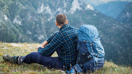 A male hiker with a backpack behind him sits on a grassy hill looking at the distant mountains. He...