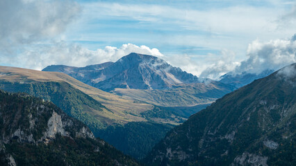 A panoramic view of a mountain range with a valley below. The mountains are covered in green vegetation and the valley is filled with lush forests. Clouds cover the peaks of the mountains.