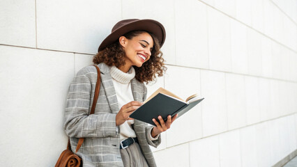 Obraz premium woman in stylish outfit and hat leans against wall, smiling while reading book