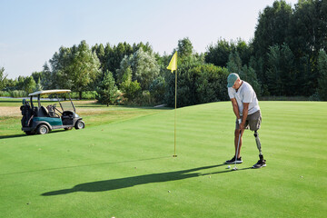 A golfer with a prosthetic leg prepares to putt on a well-maintained golf green while a cart waits...