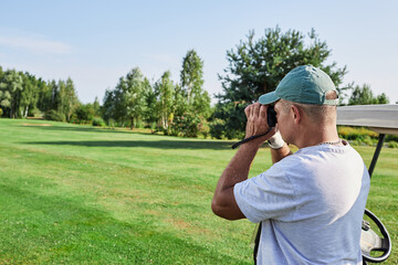 Golfer monitors the results of his shot with binoculars while standing on a green golf course under...