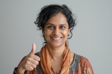 Portrait of a jovial indian woman in her 40s showing a thumb up isolated in soft gray background