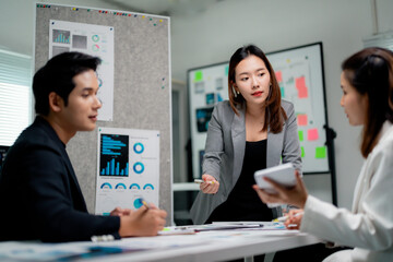 Asian businesspeople discussing during a meeting in a modern office, analyzing financial charts displayed on a wall, and using a digital tablet