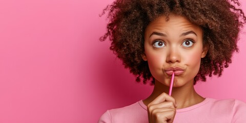 Fototapeta premium A young woman with curly hair looks thoughtfully while holding a pink pencil to her lips. The bright pink backdrop adds a playful touch to her expression of curiosity