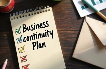 Notebook lying on wooden surface with text Business Continuity Plan. Check marks and cross marks. Graphs, envelope, pen and cup of coffee laying near. Office work planning. Risk management.