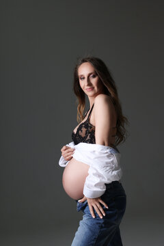 Portrait of attractive pregnant woman in shirt, jeans, with naked belly, studio photoshoot. Fashion photo of pregnancy. Young pretty girl with long hair touching her belly. Isolated, greay background